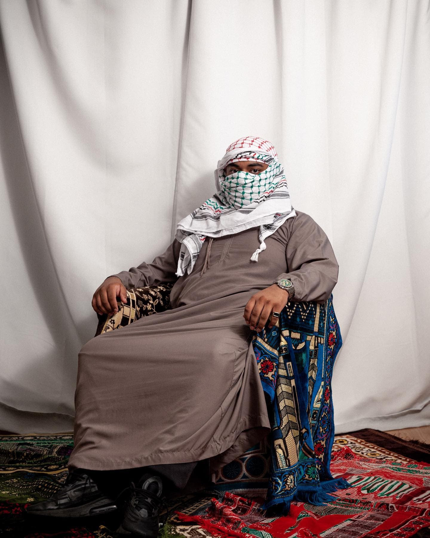 Studio portrait — seated model in a grey thobe and keffiyeh on layered prayer rugs