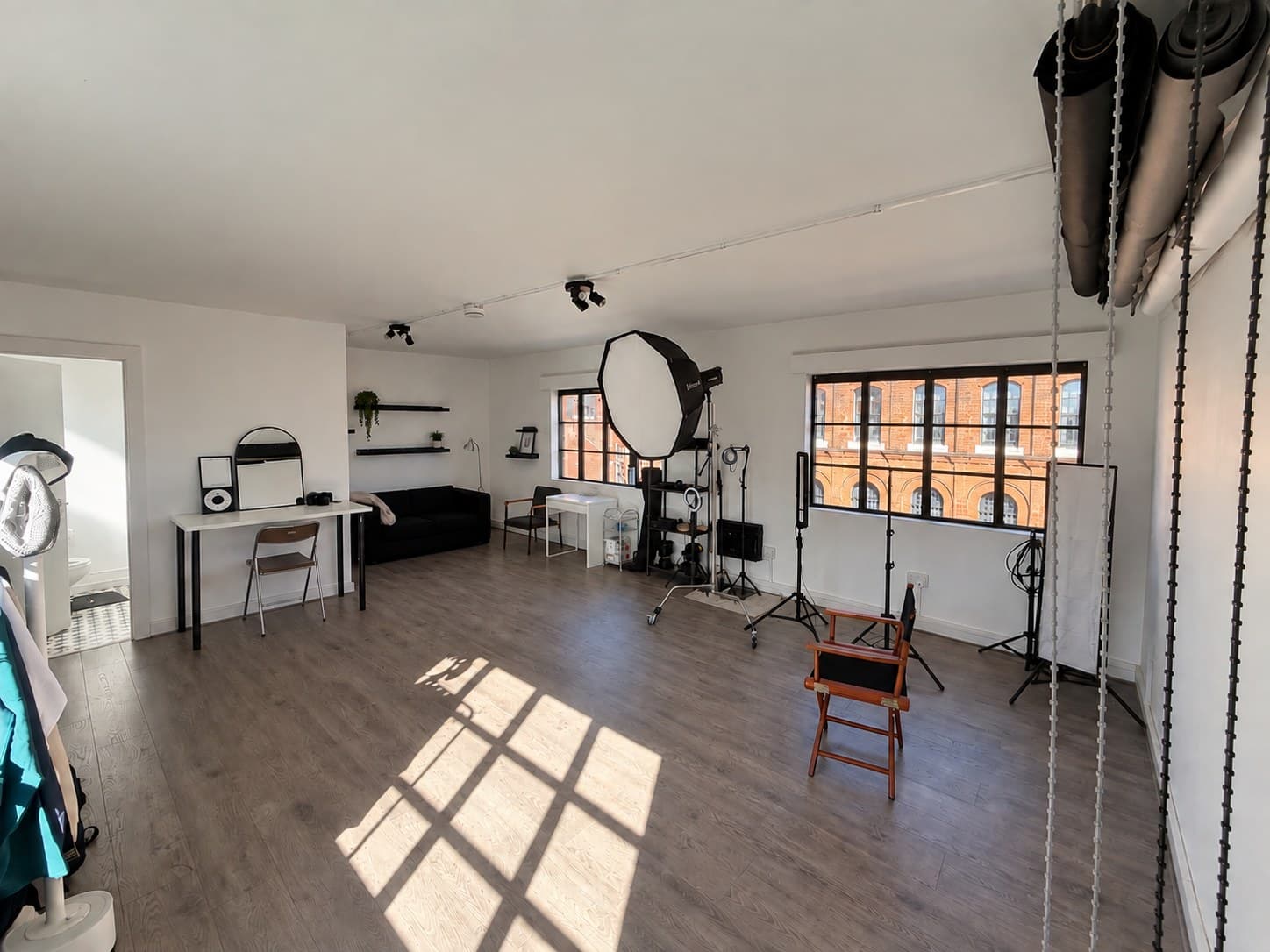 Studio One: the full room, roller backdrops racked on the wall, octabox and light stands by the windows, sofa, makeup desk and changing area